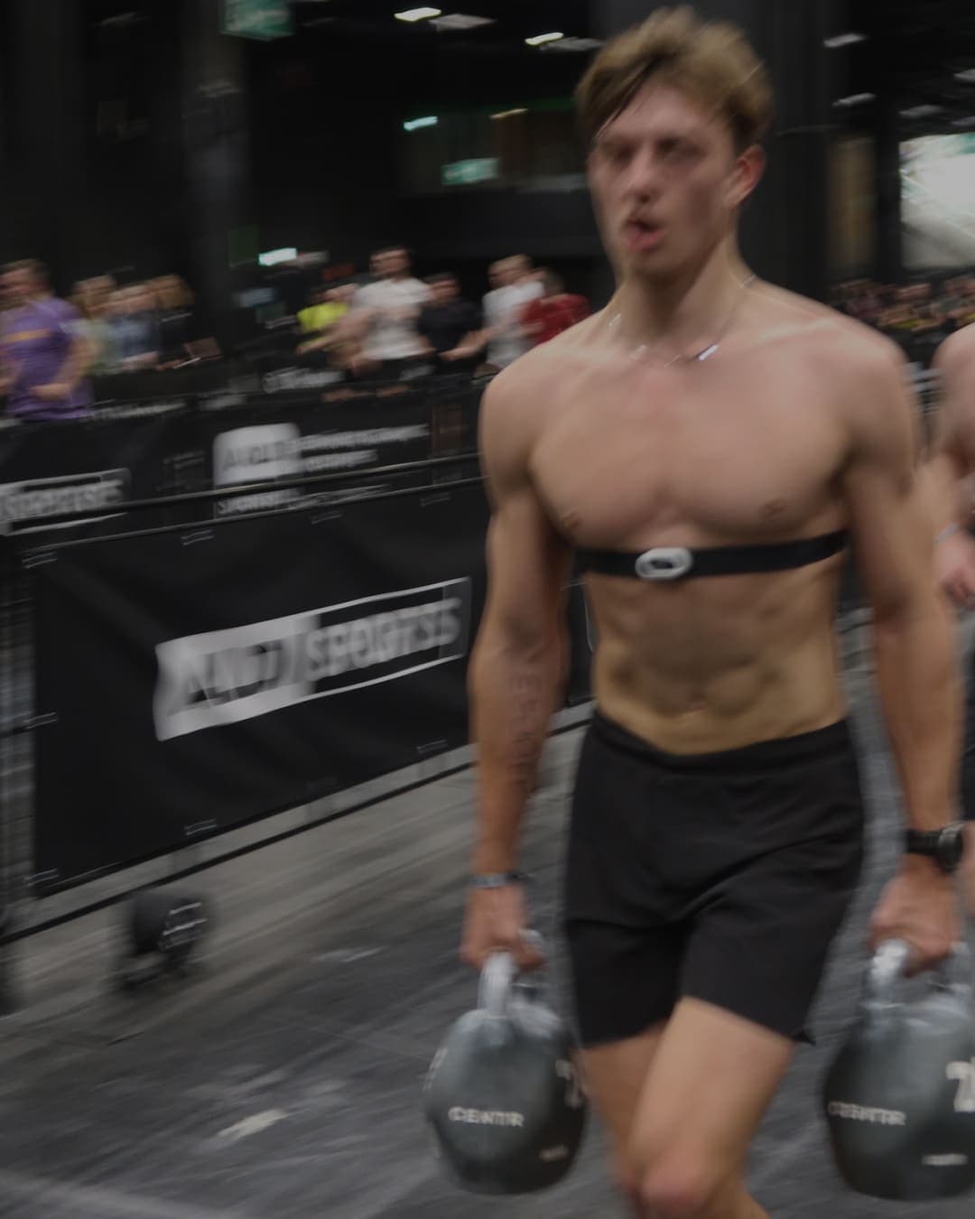Athlete in a heart rate strap carrying two kettlebells past the crowd